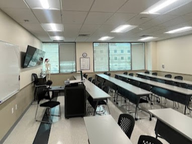 An empty classroom with rows of desks, a teacher’s station, large windows, and a wall-mounted screen.