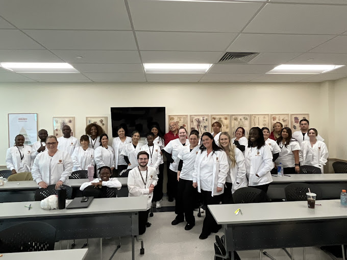 A group of nursing students in white lab coats posing together in a classroom with desks and medical anatomy posters on the wall.