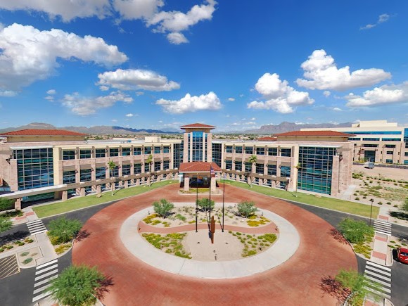 A large modern hospital complex with glass-front buildings surrounding a circular driveway and landscaped courtyard under a bright blue sky.