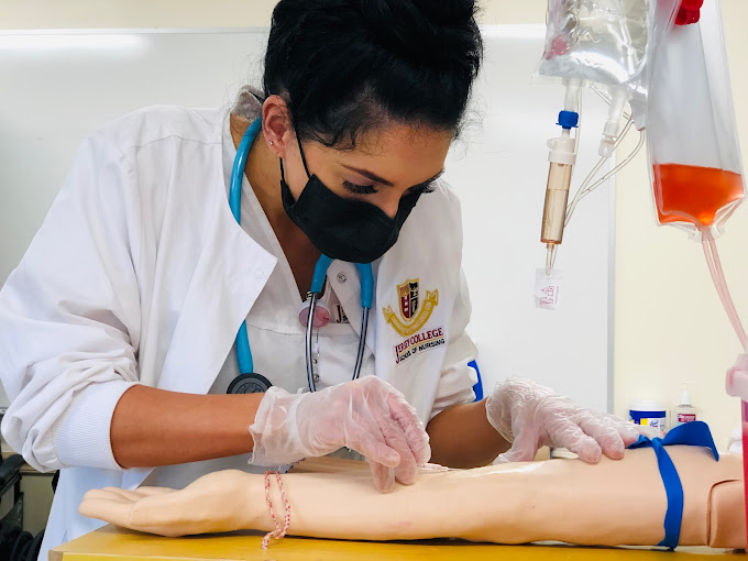 A nursing student wearing gloves and a mask practices starting an IV on a training arm with IV bags hanging beside her.